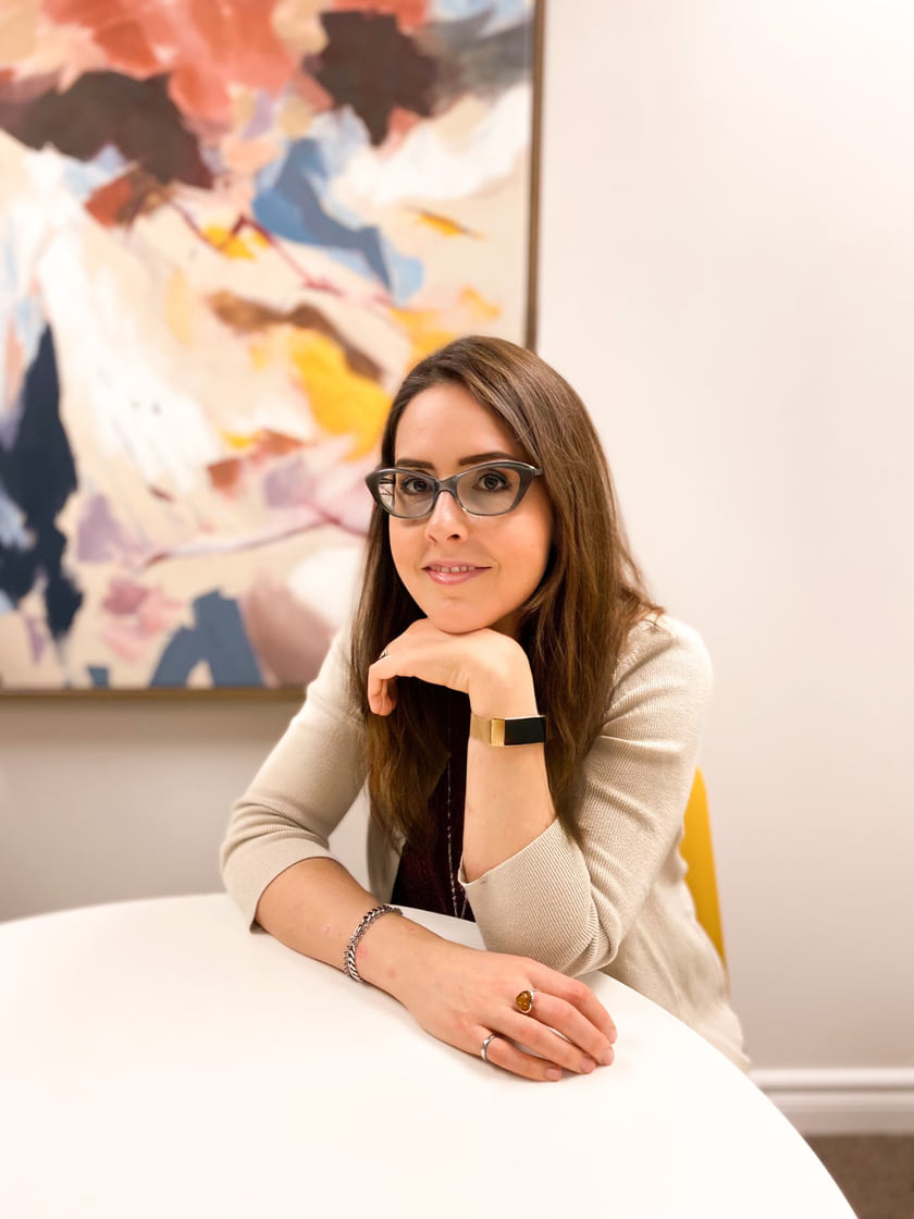 Marika Fradette seated at a white table, resting her chin on her hand, in front of a large abstract painting.